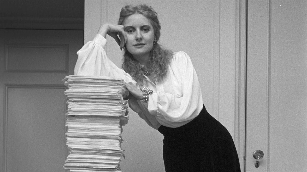A woman in business clothing leans on a large stac