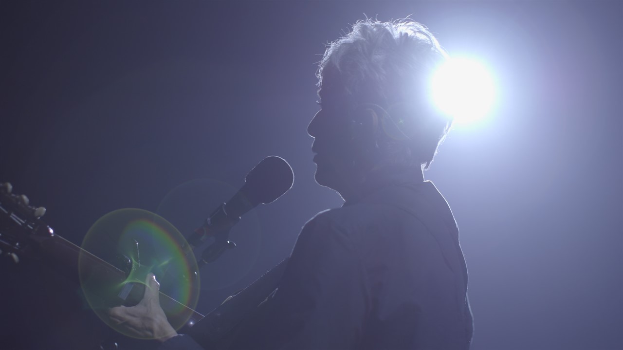 Joan Baez, backlit, sings at a microphone playing 
