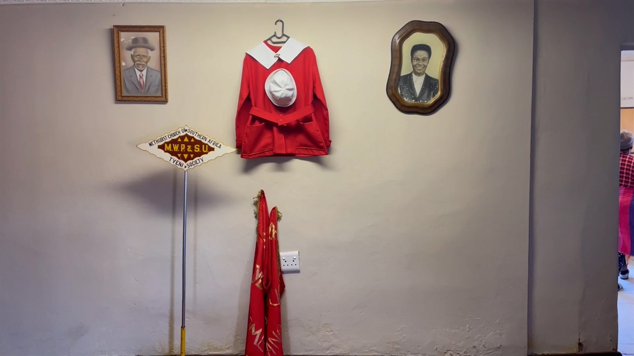 A small, red jacket and white hat hang on a wall