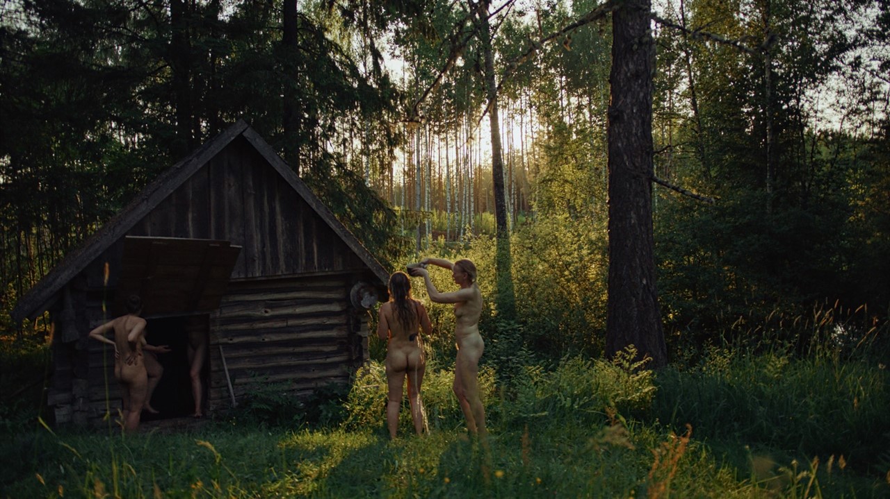 Two naked woman stand outside of a wooden sauna