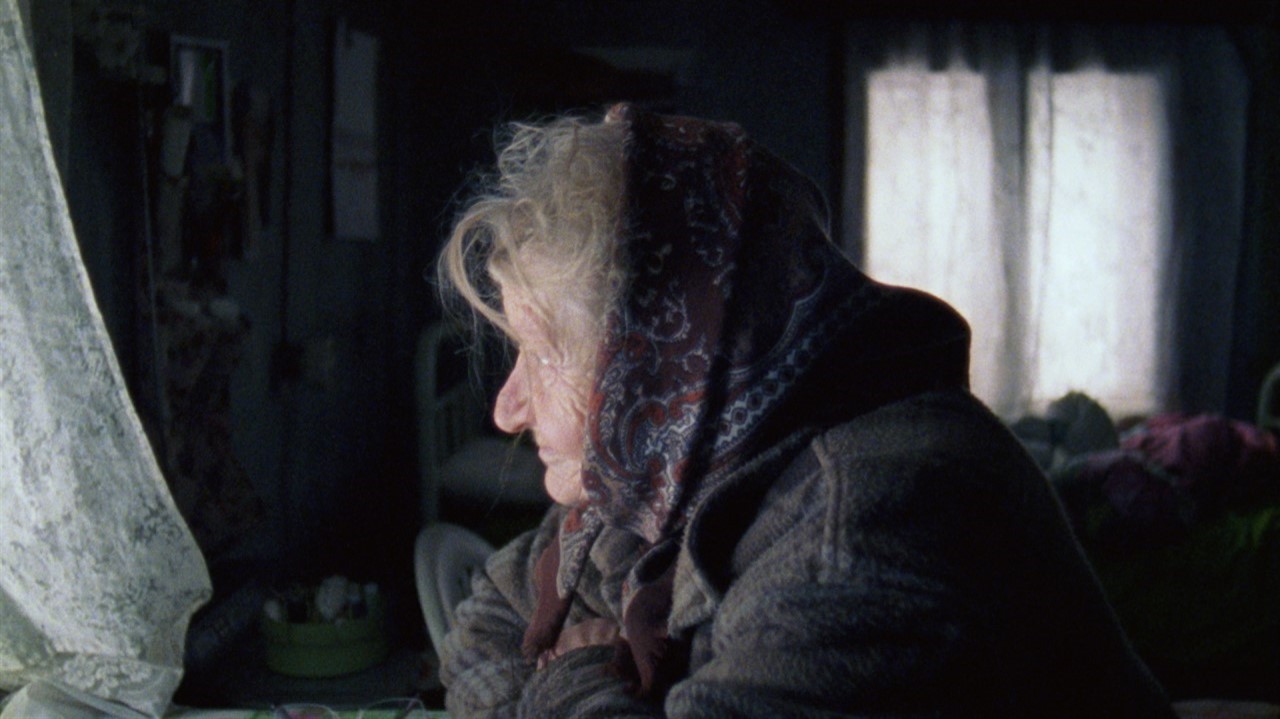 An elderly woman in a headscarf looks out a window